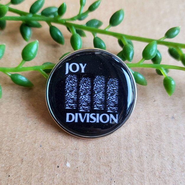 Round black badge with four narrow repetitions of the Unknown Pleasures chart between JOY and DIVISION