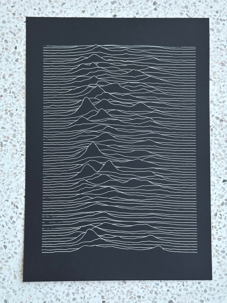 A top-down photograph of a white-ink Unknown Pleasures-inspired pen plot on black paper